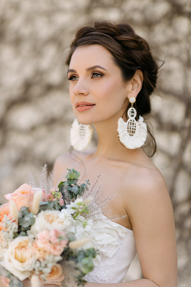 Bride in Montreal with a delicate braided updo, gazing off in the distance. Her wedding makeup is soft and natural, complemented by statement earrings and a bouquet featuring blush tones, perfect for a romantic bridal look