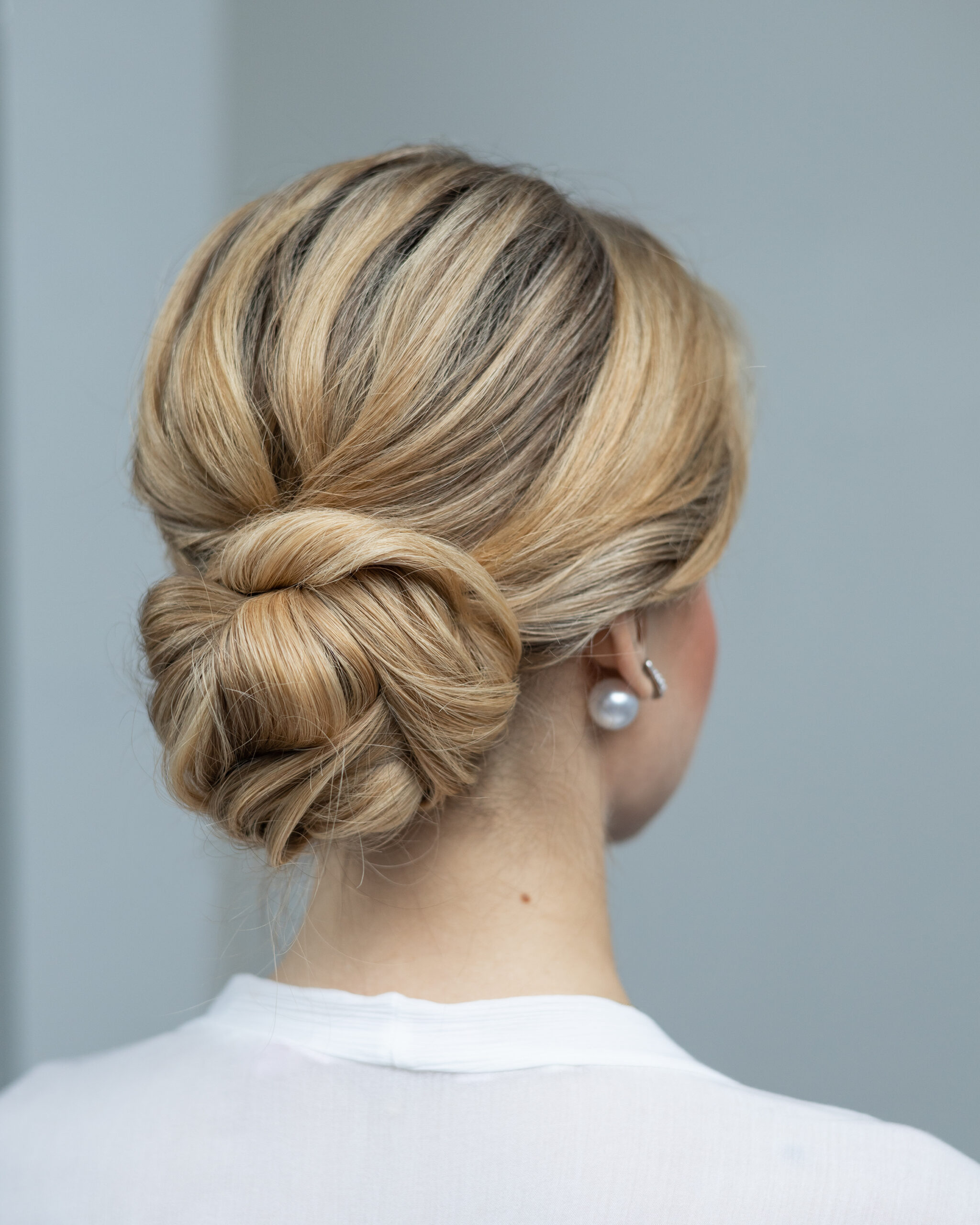 A sophisticated bridal hairstyle featuring a twisted low bun with blonde highlights, perfect for a chic Montreal wedding, paired with classic pearl earrings for a touch of grace