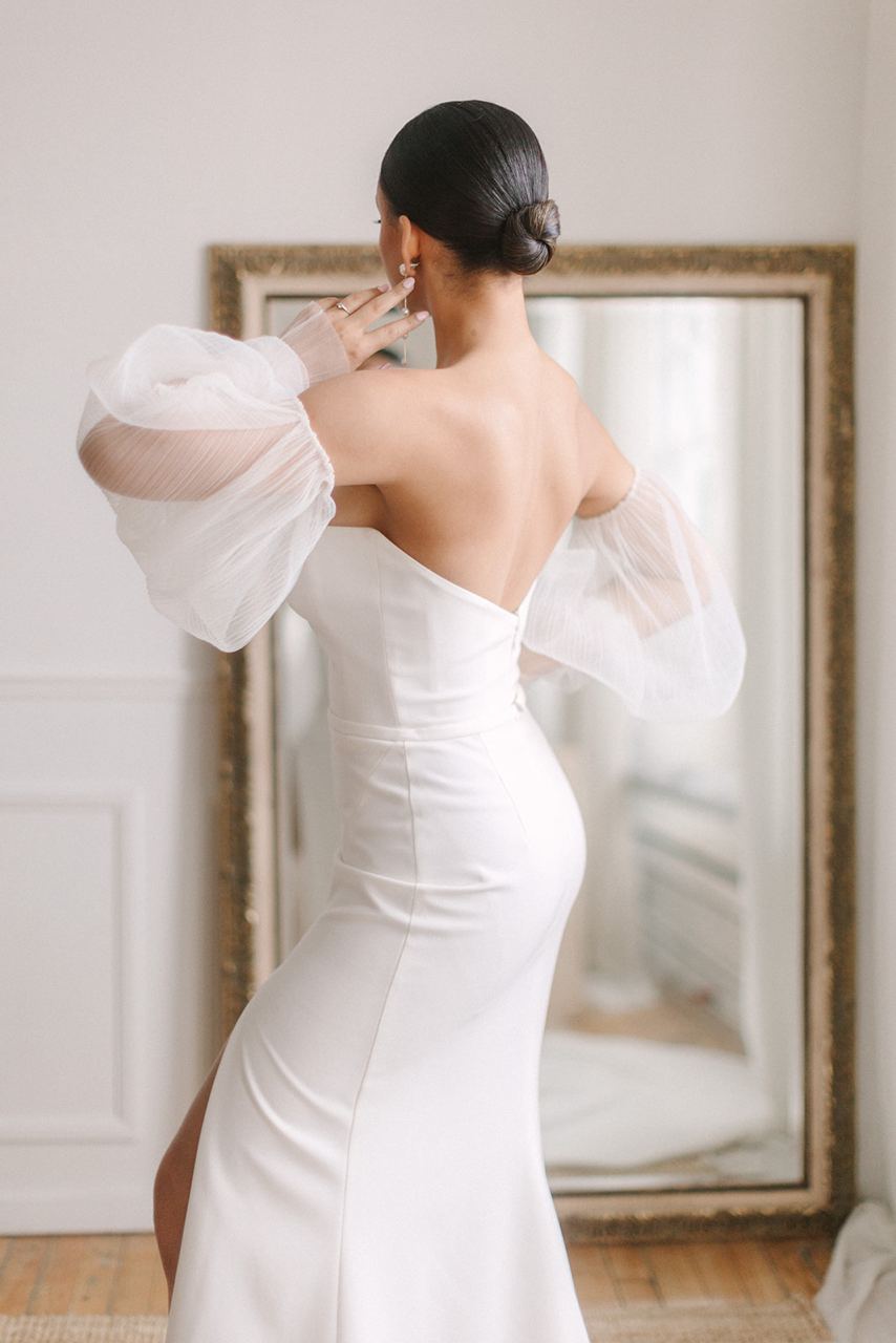 An elegant bride in a chic wedding dress admiring her reflection in the mirror, featuring a classic updo with a sleek bun crafted by a Montreal bridal hairstylist from Beauty Trend.