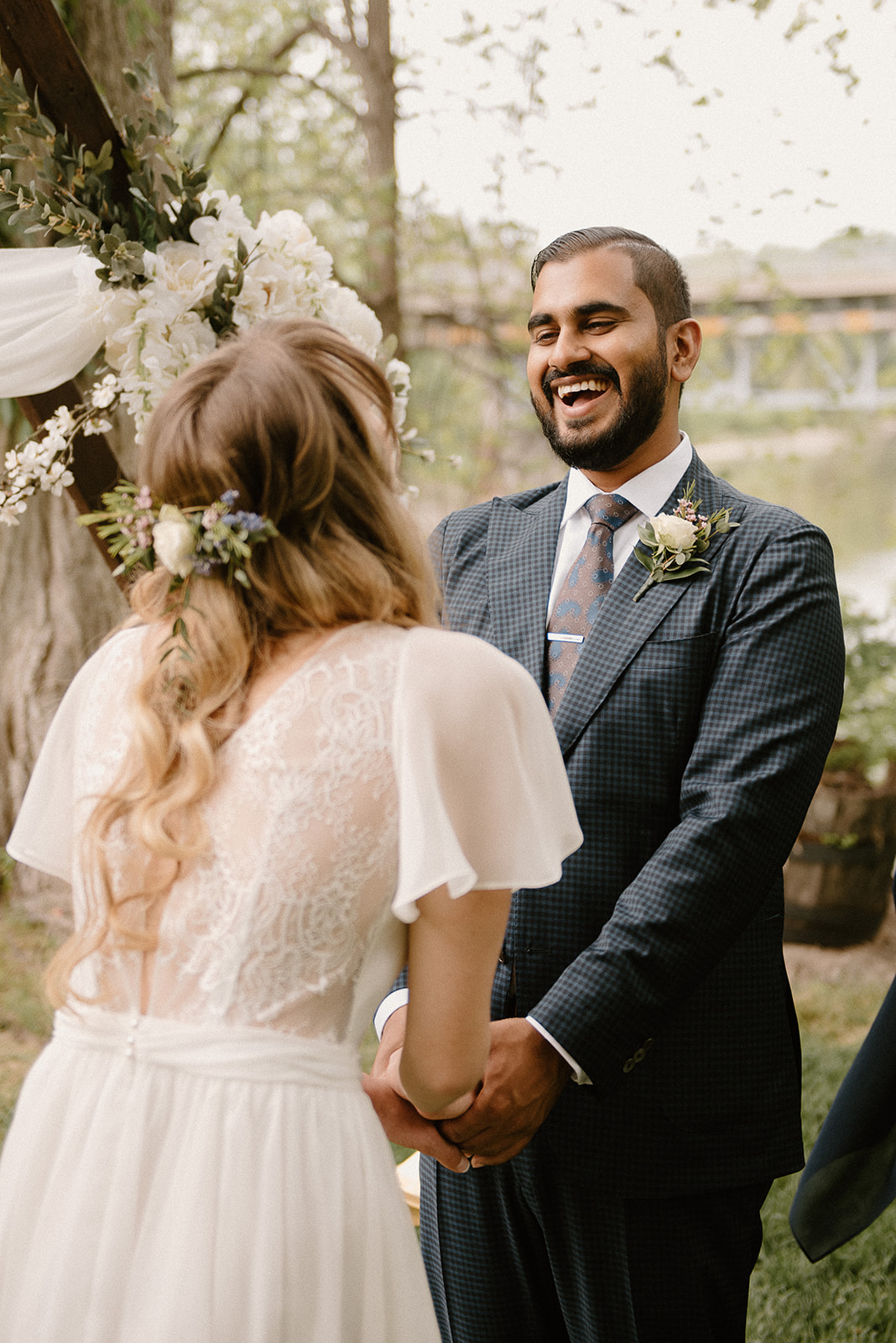 Joyful bridal couple exchanging vows under a floral arch in Montreal, with the bride's intricate lace dress and natural hairstyle highlighted by the soft daylight.