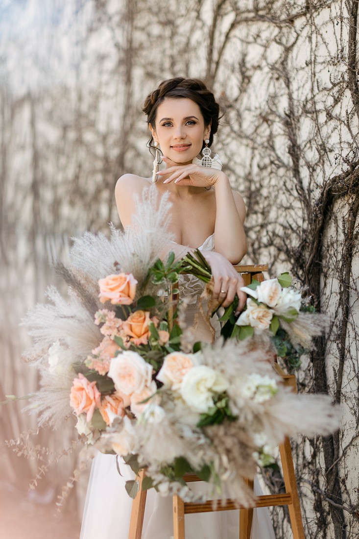 Bride in Montreal with a whimsical wedding hairstyle, posing against a textured backdrop, holding a lush bouquet with peach and cream roses, embodying a bohemian beauty trend