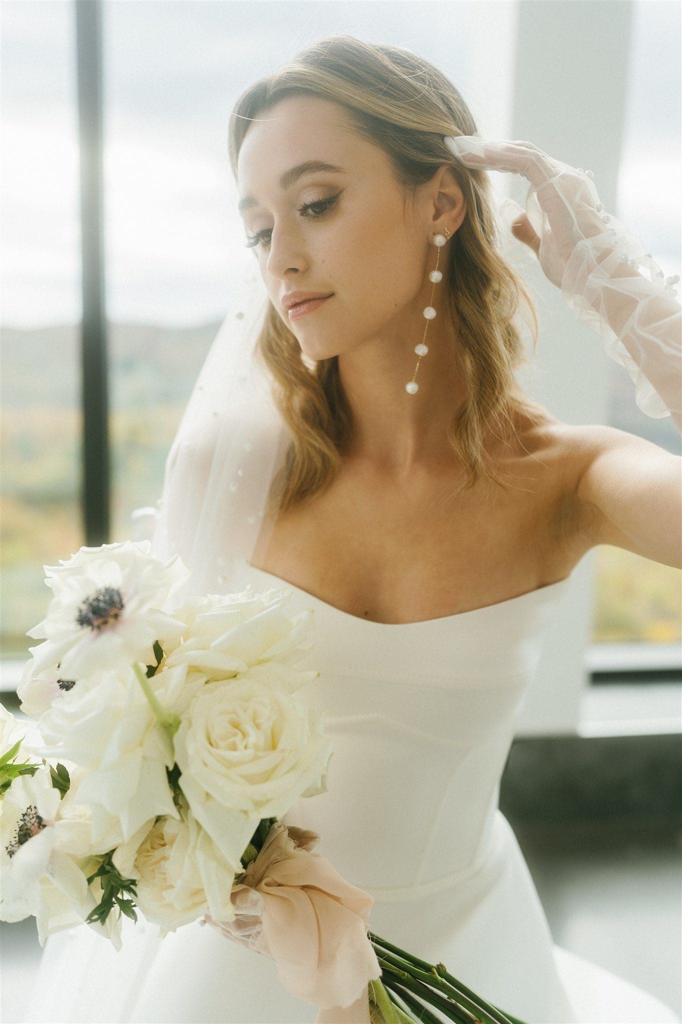 A bride in Montreal holding a classic white bouquet, styled by bridal hair and makeup experts with pearl drop earrings and a soft wavy hairstyle.