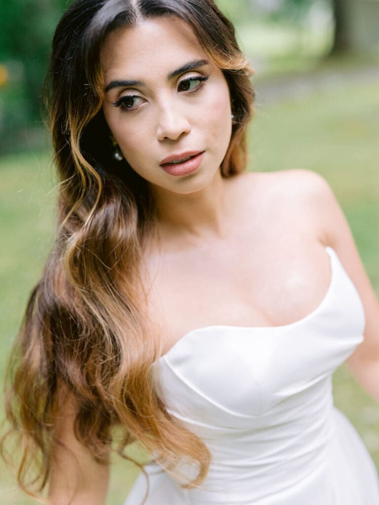 A bride in Montreal with loose romantic waves, natural bridal makeup, and a strapless white gown, posing in a tranquil garden setting.